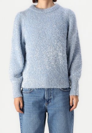Personne portant un pull en peluche bleu clair associé à un jeans bleu taille haute et ample, se tenant devant un fond blanc uni.
