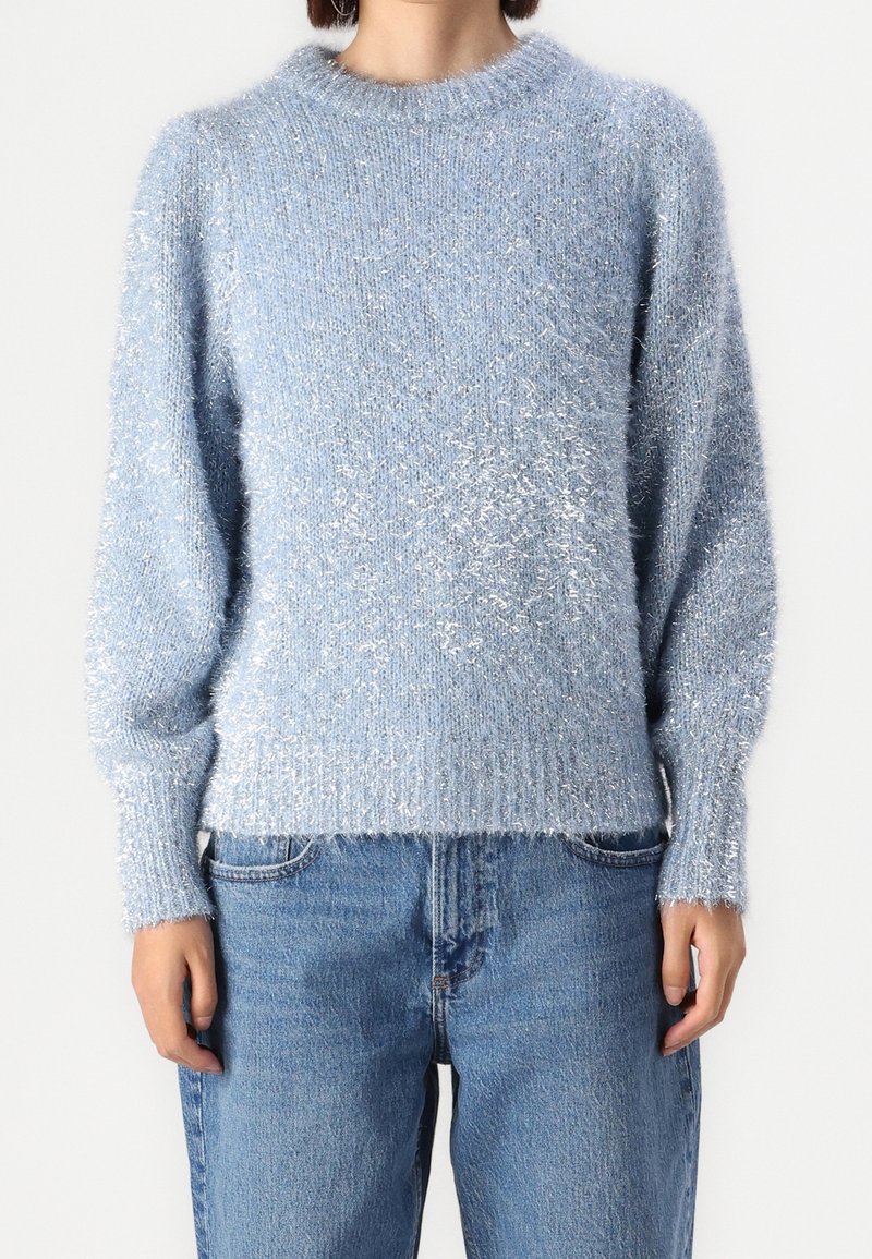 Eine Person trägt einen flauschigen hellblauen Pullover, kombiniert mit hochgeschnittenen, locker sitzenden blauen Jeans, und steht vor einem schlichten weißen Hintergrund.