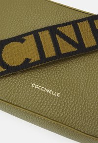 Olivově zelená texturovaná kožená taška s černým a hořčicovým pleteným popruhem, který zdobí výrazné písmena a zlaté logo "COCCINELLE" vpředu.