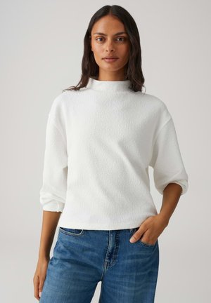 Witte, gestructureerde trui met een hoge kraag, doorgestikte schouders en lange, opgerolde mouwen. Gecombineerd met blauwe spijkerbroek.