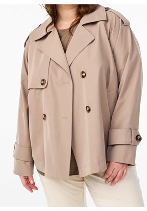 Personne portant un trench-coat beige double boutonnage avec des boutons noirs et un pantalon beige assorti sur fond blanc.
