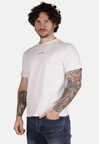 T-shirt bianca in cotone con maniche corte, caratterizzata da un logo viola "PROPAGANDA" sul petto. Il modello indossa jeans e ha tatuaggi visibili.