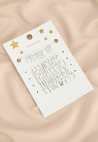 Strieborná súprava písmeniek na bielej kartičke so zlatými hviezdami. Text znie "Charm Me," obsahuje písmená A-Z, pripevnené malými sponkami.