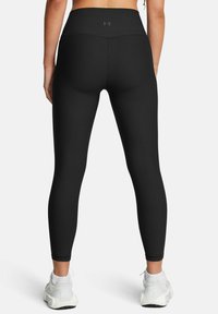 Leggings nere a coste con vita alta, dotate di un piccolo logo, abbinate a scarpe da ginnastica bianche su uno sfondo neutro.