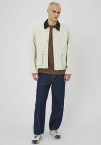 Veste crème avec col marron, poches ; chemise marron en dessous, pantalon en denim bleu foncé et baskets claires. Design minimaliste, coupe décontractée.