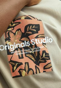 Bežinis marškinėlis su koraliniais ir juodais grafikais, vaizduojančiais lapų raštus, ir baltais užrašais "Originals Studio" bei prekės ženklo informacija.