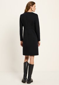 Frau in einem enganliegenden schwarzen knielangen Kleid mit langen Ärmeln, mit dem Rücken zur Kamera gewandt, kombiniert mit schwarzen kniehohen Lederstiefeln und goldenen Creolen.