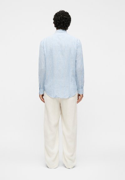 Άνδρας με σγουρά μαλλιά, φορώντας μπλε και άσπρη ριγέ μακρυμάνικη μπλούζα και φαρδιά μπεζ παντελόνια, με την πλάτη σε ένα απλό φόντο.