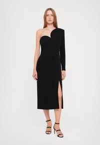 Crna, uska midi haljina s jednim naramenicom, asimetričnog dizajna, bočnim prorezom i glatkom teksturom tkanine. Prozirne potpetice dopunjuju izgled.