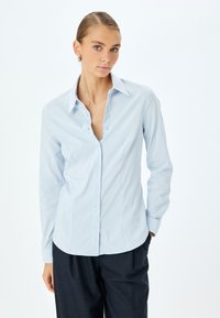 Camicia a maniche lunghe azzurro chiaro con colletto appuntito, chiusura a bottoni frontale e design aderente, abbinata a pantaloni scuri. Tessuto con texture liscia.