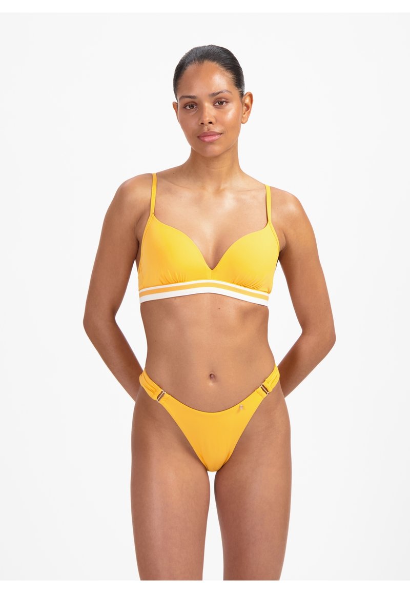 Frau mit dunklem Haar in einem gelben zweiteiligen Badeanzug mit weiß gestreiftem Band, die mit den Händen hinter dem Rücken vor einem einfarbigen Hintergrund steht.