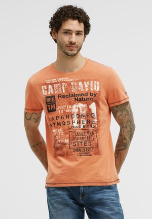Man with curly hair wearing an orange graphic T-shirt and blue jeans, standing with hands behind his back against a plain background.