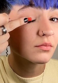 Een persoon houdt een kleine rode make-up applicator tegen hun gesloten ooglid. Ze hebben blauw haar en dragen een lichte geribde trui.