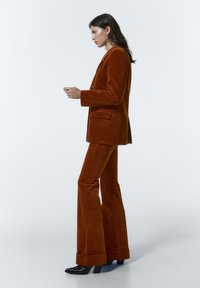 Costume en velours côtelé rouille avec un blazer cintré et un pantalon évasé, présentant une texture lisse et des bottes noires à talons. Design simple et élégant.