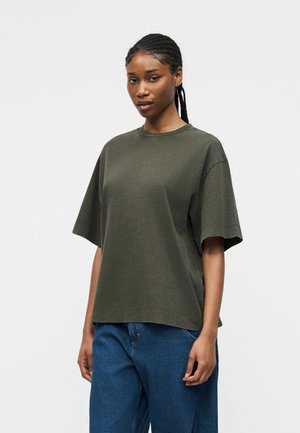 Ženska s pletenicami, oblečena v preveliko olivno zeleno majico in modre kavbojke, stoji pred enobarvnim svetlo sivim ozadjem.