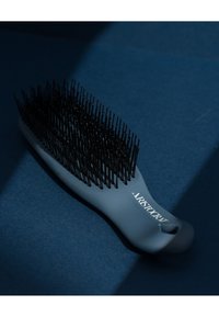 Grå plast hårbørste med et bredt håndtak og svarte børster arrangert i et buet mønster; har teksten "ARISTOCRAT" på håndtaket.