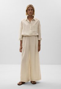 Zweiteiliges Outfit aus Leinen in Creme, bestehend aus einem knöpfbaren Hemd mit Tasche und weit geschnittenen Hosen, beide mit dezenten vertikalen Streifen.
