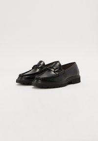 Mocassins en cuir noir avec un design texturé, présentant un dessus cousu, des accents métalliques et une semelle en caoutchouc épaisse.