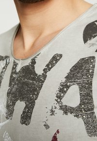 Camiseta gris con escote en V, que presenta un estampado desgastado y detalles de tachuelas en la parte frontal. La tela parece suave y ligera.