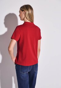 Polo rouge à manches courtes, en coton. Présente un col classique et une texture lisse. Porté avec un jean en denim.