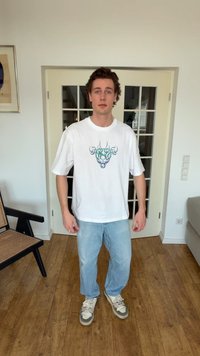 Ung mann iført hvit t-skjorte med grafisk trykk, lyseblå jeans og joggesko, står i et rom med tregulv og glassdører bak seg.