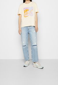 Mujer vestida con una camiseta color crema con la impresión "Paz y Amor", jeans desgastados de color azul claro y zapatillas en tonos pastel, de pie contra una pared blanca.