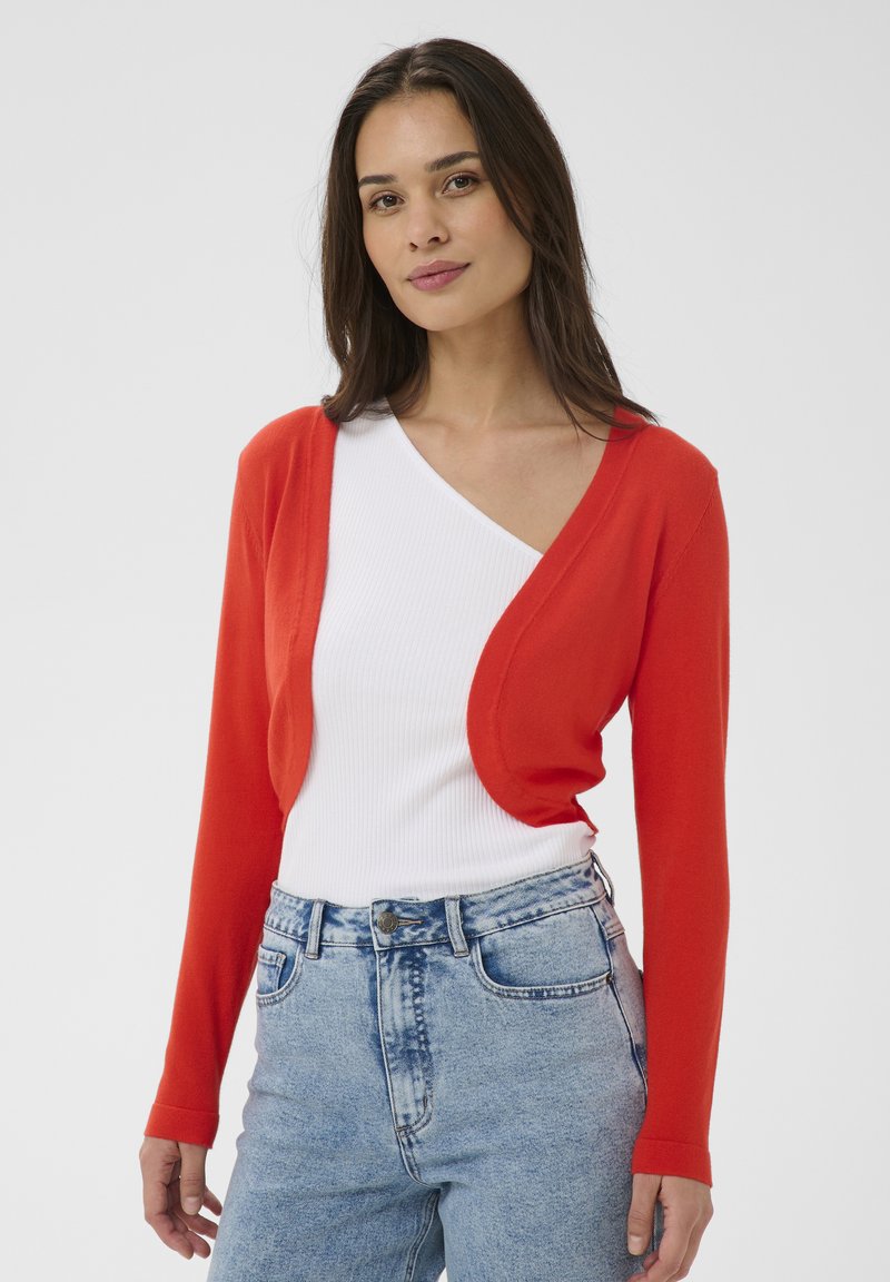 Femme portant un cardigan rouge cropé par-dessus un haut blanc sans manches et un jean taille haute bleu clair, debout devant un fond uni.