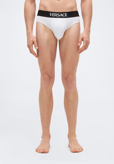 Modèle masculin portant un slip blanc avec une ceinture noire ornée du logo Versace, debout pieds nus devant un fond uni.