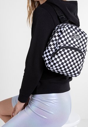 Mochila a cuadros en blanco y negro hecha de tela, con un bolsillo frontal con cremallera y correas ajustables, llevada sobre una sudadera negra con capucha.