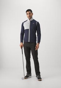 Námornícky modrá a biela golfová bunda na zips s vysokým golierom; textúrovaná látka má dizajn s farebnými blokmi. Čierne nohavice a šedé golfové topánky dokončujú vzhľad.