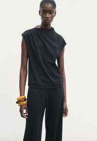 Black short-sleeve top with a high neck, paired with wide-leg black pants. A stack of orange and green bangles adorns the left wrist.