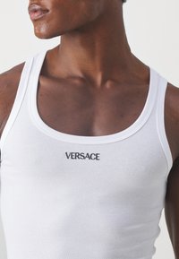Valge ribastatud tank top, millel on ümar kaelus ja keskel väike must "VERSACE" logo. Kujundatud istuvaks ilme saavutamiseks kergest kangast.