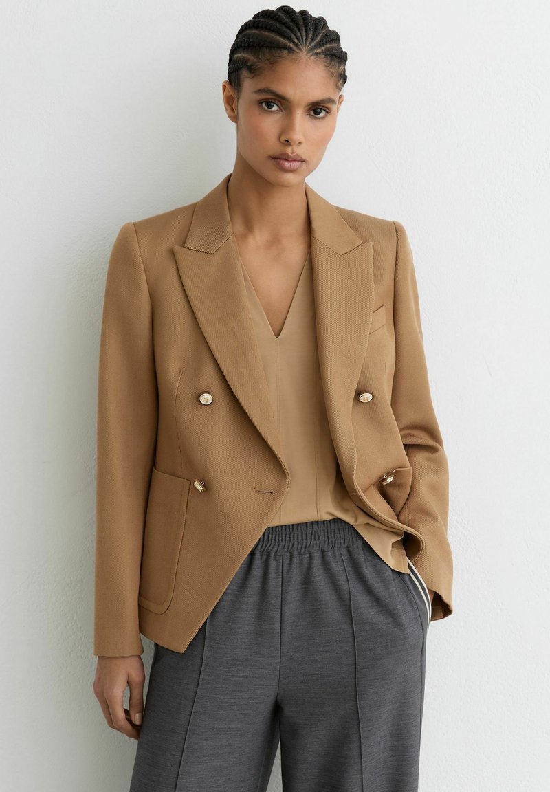 Tan dobbeltradet blazer i tekstureret stof med gulddetaljer, kombineret med en beige top og grå bukser med elastisk talje.