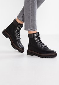 Bottes en cuir noir montant à la cheville, avec un bout arrondi, des œillets en argent pour les lacets, et une semelle en caoutchouc présentant un motif de bande de roulement robuste.