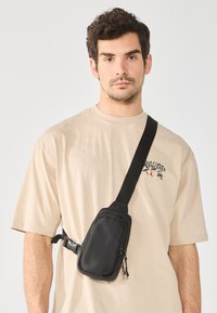 Borsa a tracolla nera in tessuto con scomparti con zip e cinturino regolabile, indossata a tracolla su una t-shirt beige chiaro con un design grafico.