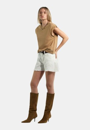 Donna con top marrone senza maniche, pantaloncini bianchi strutturati con cintura nera e stivali marroni in camoscio al ginocchio, su sfondo chiaro uniforme.