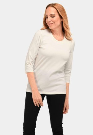Haut blanc à manches longues avec un col rond et des détails subtils. Porté avec un pantalon noir, mettant en valeur une coupe décontractée et une texture douce.