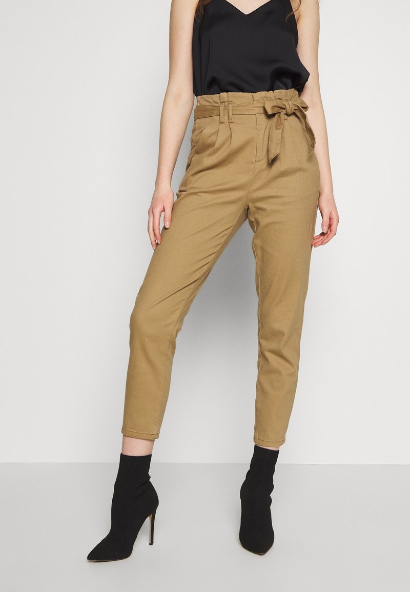 Béžové kalhoty vysokého pasu s detailem opasku, zúženými nohavicemi a měkkým bavlněným materiálem. Nosit s černými přiléhavými kotníkovými botami.