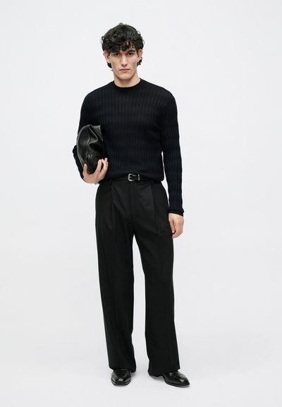 Μαύρο πουλόβερ με υφή και μοτίβο ρόμβων, συνδυασμένο με μαύρο παντελόνι και παπούτσια. Το μοντέλο κρατά μια μαύρη δερμάτινη τσάντα.