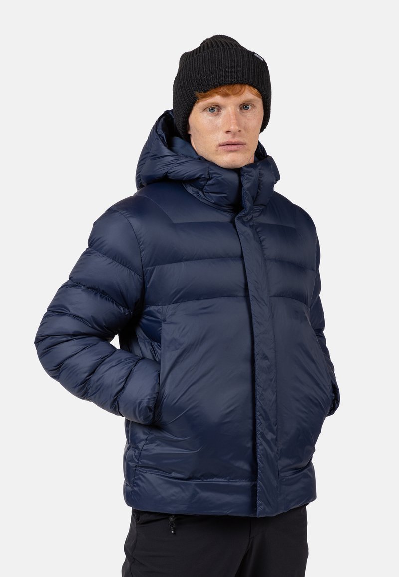 Giacca a vento in blu scuro, con cappuccio, design trapuntato, tasche frontali e materiale sintetico lucido. Abbinata a un berretto in maglia nero.