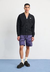 Svart cardigan med knappar och en Nike-logotyp, matchad med lila tie-dye denimshorts, vita strumpor och svarta skor. Enkelt bakgrund.