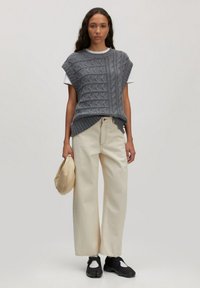 Grijze kabelgebreide mouwloze trui over een wit shirt, gecombineerd met hoge crèmekleurige wijde broek en zwarte schoenen, met een beige tas.