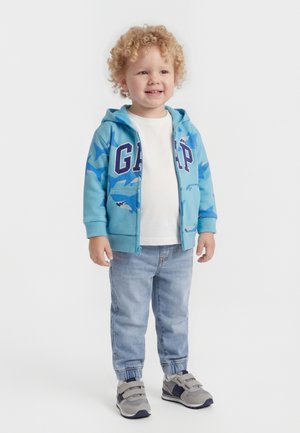 Νεαρό παιδί με σγουρά μαλλιά φοράει μπλε φούτερ GAP με σχέδιο καρχαρία, λευκό πουκάμισο, ανοιχτόχρωμα τζιν και γκρι αθλητικά παπούτσια, στέκεται χαμογελώντας.