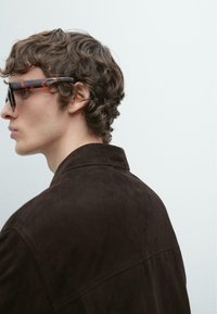 Giacca in camoscio marrone con collo a punta, indossata sopra una camicia strutturata. Modello con occhiali da sole tartarugati, con capelli scuri e ondulati.