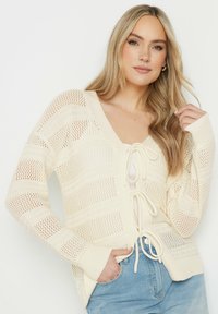 Krämfärgad cardigan med djup V-ringning, öppna stickade ränder och knytframslutning. Lång ärm med ribbade manschetter, bärs över denimshorts.
