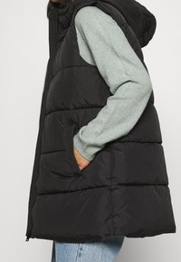 Svart vadderad väst med huva, försedd med dragkedja och två sidofickor. Texturen är slät med ett quiltat mönster.