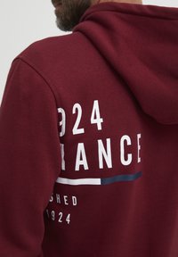 Burgunderhette med en hvit grafikk som viser "924" og teksten "FRANCE", horisontale striper i marineblått og hvitt, samt en strukturt stofffinish.