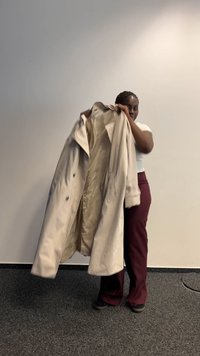 Beige Trenchcoat mit einer glatten Textur, großen Revers und Knopfverschlüssen, gehalten von einer Person, die ein weißes Oberteil und bordeauxfarbene Hosen trägt.