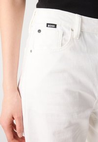 Gros plan d'une personne portant un jean blanc avec une étiquette noire "BOSS" sur la poche avant et un haut noir.