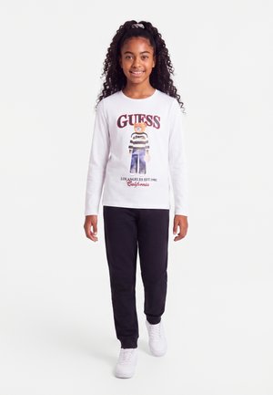 Момиче с къдрава коса, носещо бяла блуза с дълъг ръкав Guess с графика на мечка и черни панталони, върви напред на бял фон.